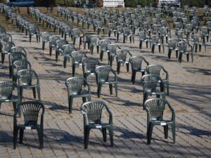 An outdoor setting with rows of empty green plastic chairs arranged on a paved area, creating a pattern.